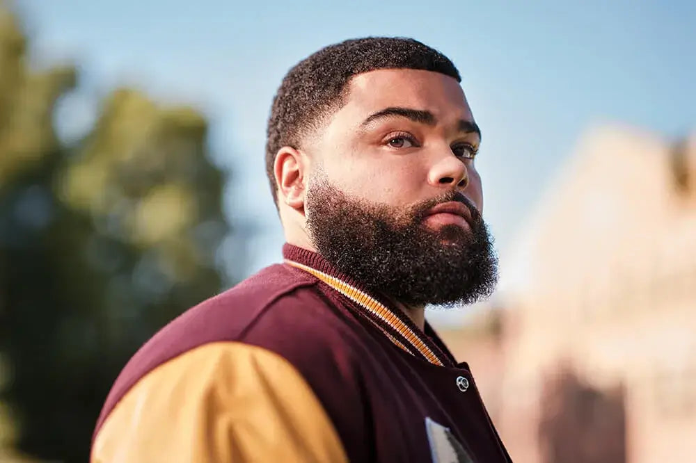 Discover Textured Hair Styling + Haircare recommendations from American Crew. Model seen smiling confidently facing the camera outdoors on a college campus while wearing a burgundy varsity jacket with yellow sleeves.