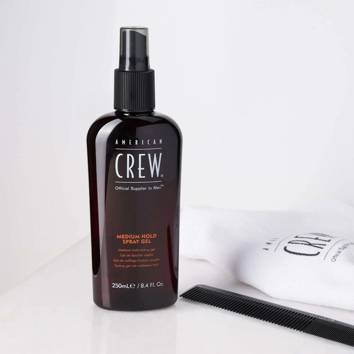 American Crew Medium Hold Spray Gel next to a branded white towel and black comb on a white table